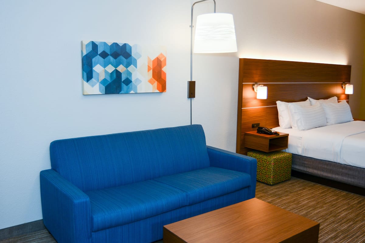 A hotel room with a blue sofa, a wooden coffee table, a bed with white linens, a nightstand with a lamp, and a geometric wall art piece.