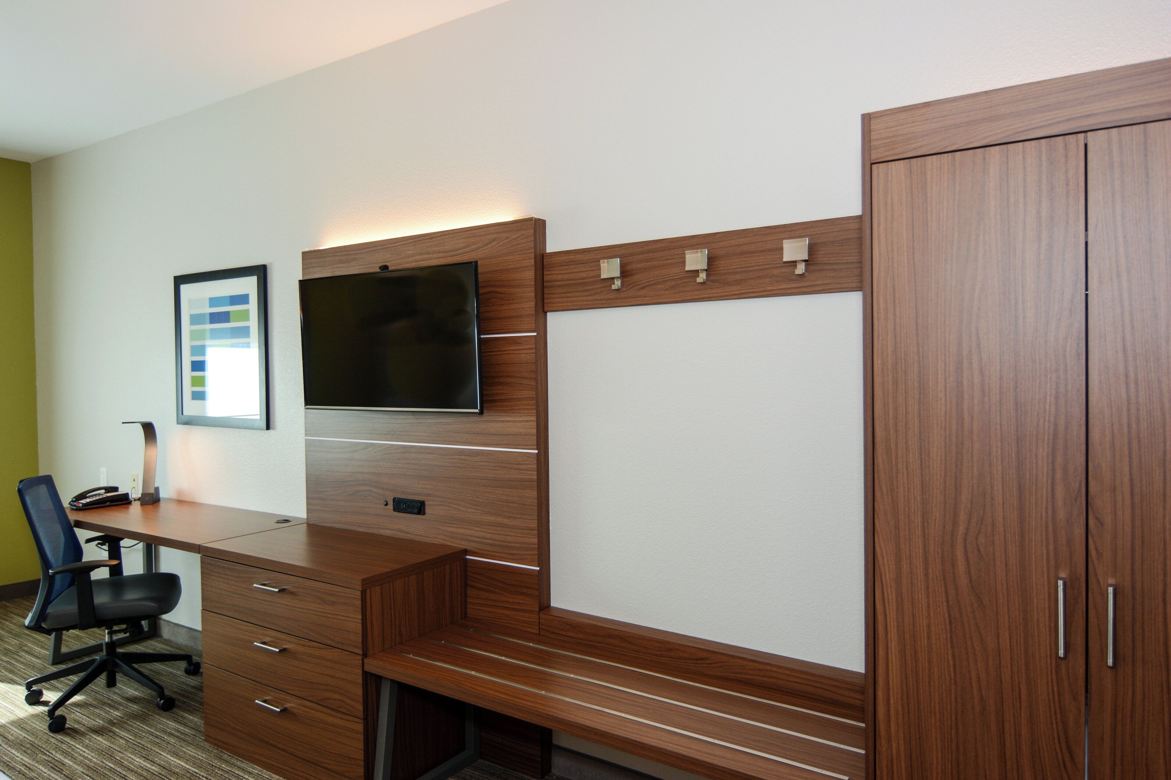 A hotel room with a wooden desk, a flat-screen TV, a chair, and a wardrobe.