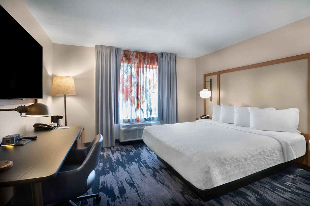 A hotel room with a large bed, a desk with a lamp, a flat-screen TV, and a window with curtains.