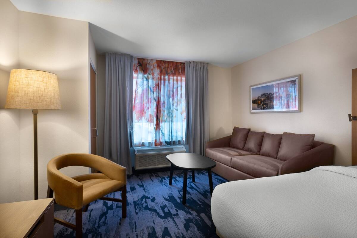 A hotel room with a sofa, a small table, a chair, a lamp, and a window with curtains.