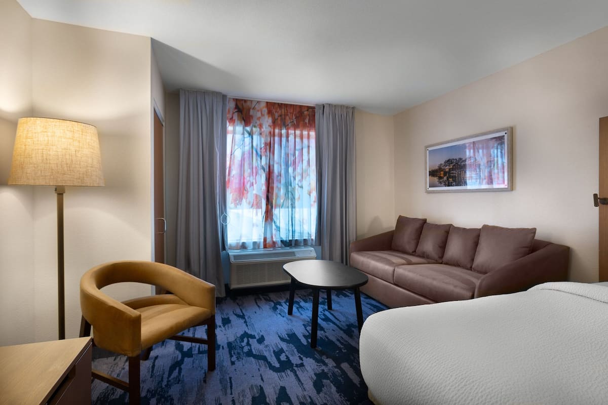 Une chambre d’hôtel avec un canapé, une petite table, une chaise, une lampe et une fenêtre avec des rideaux.