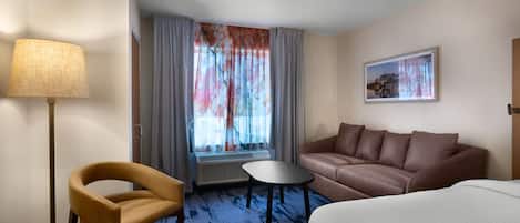 Habitación de hotel con sofá, una mesita, una silla, lámpara y ventana con cortinas.