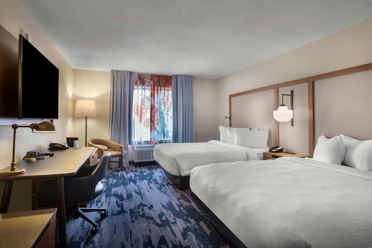 Une chambre d’hôtel avec deux lits, un bureau, une chaise, une télévision et une fenêtre avec des rideaux.