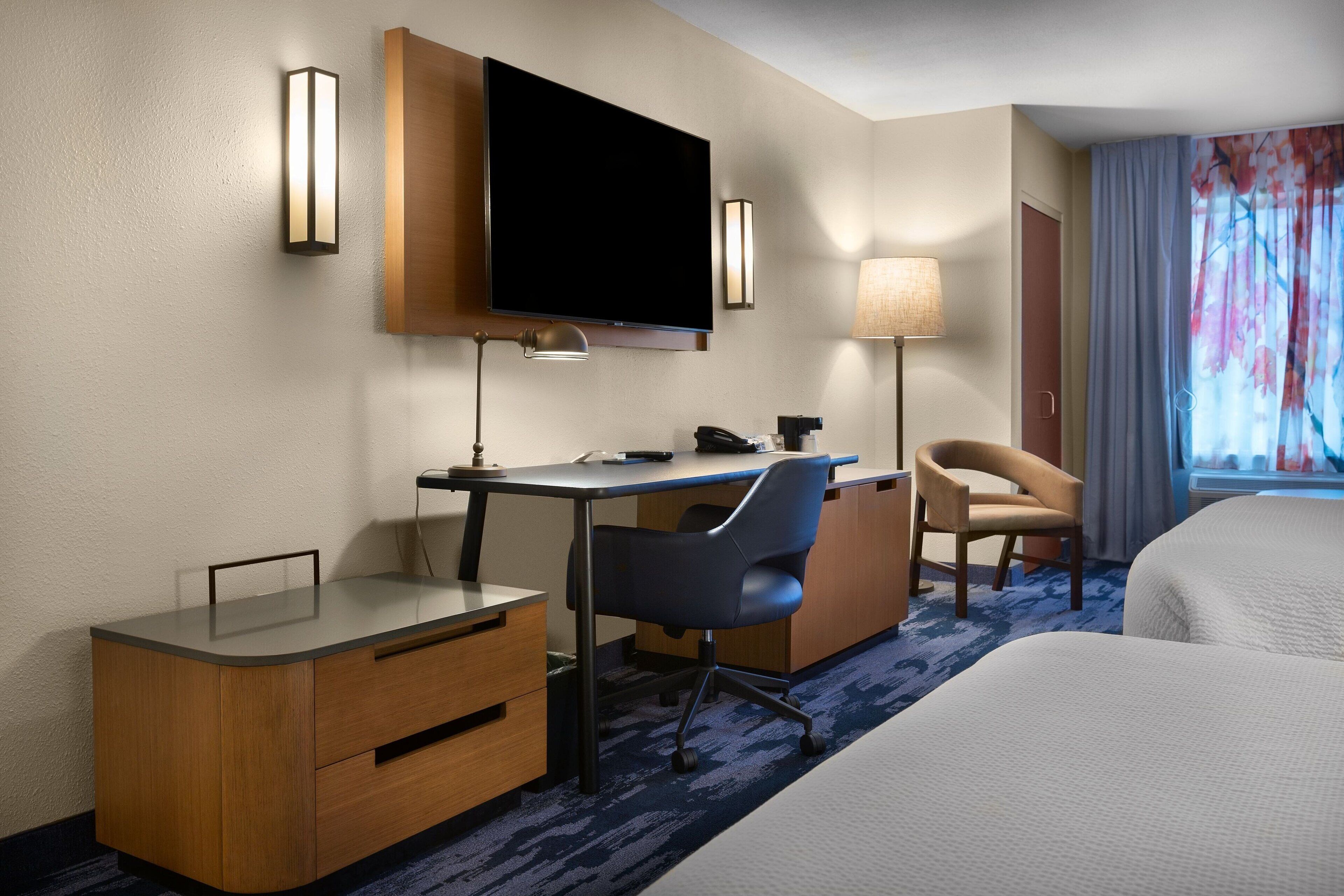A hotel room with a bed, a desk with a chair, a television, a lamp, and a window with curtains.