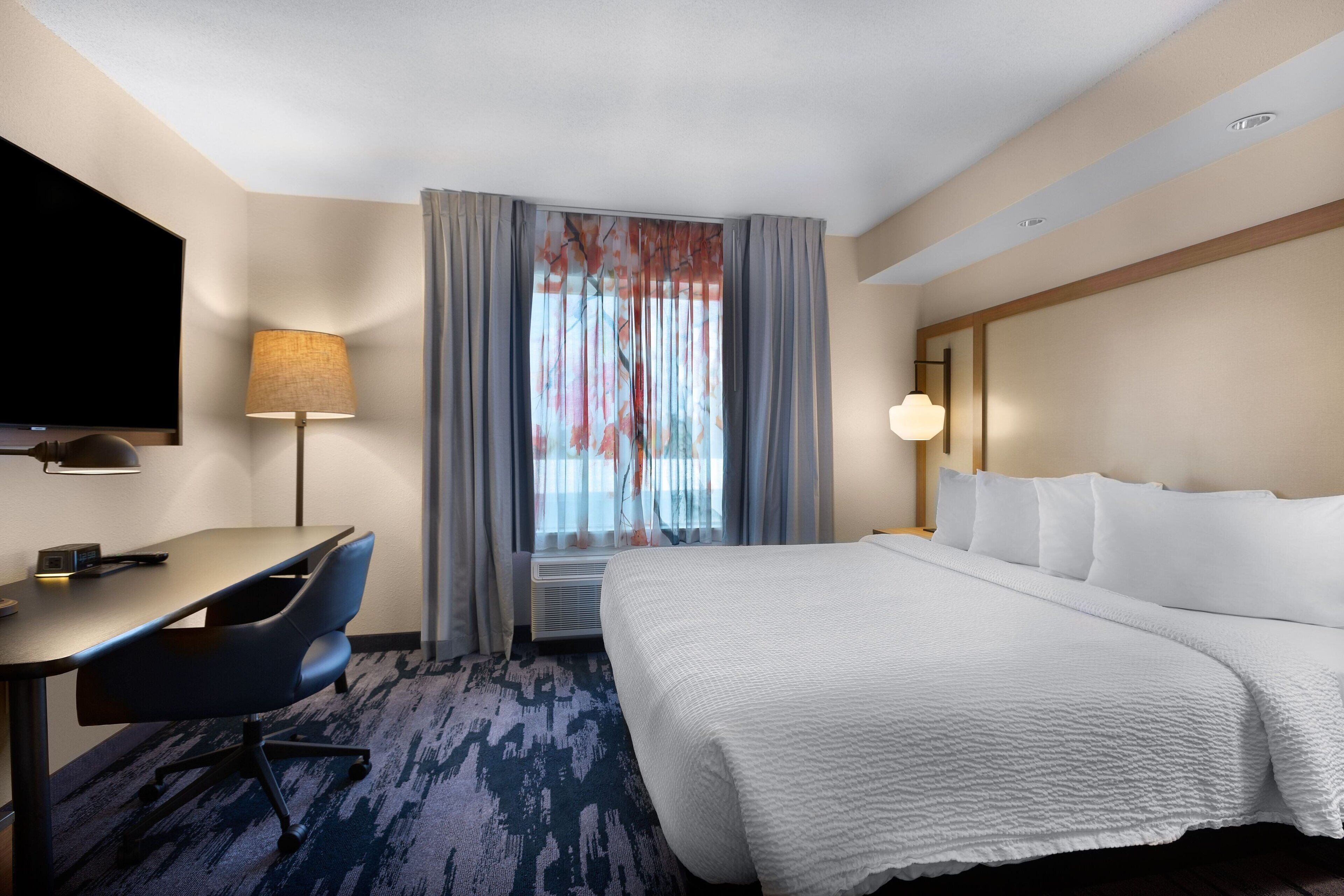 A hotel room with a bed, a desk, a chair, a TV, and a window with curtains.