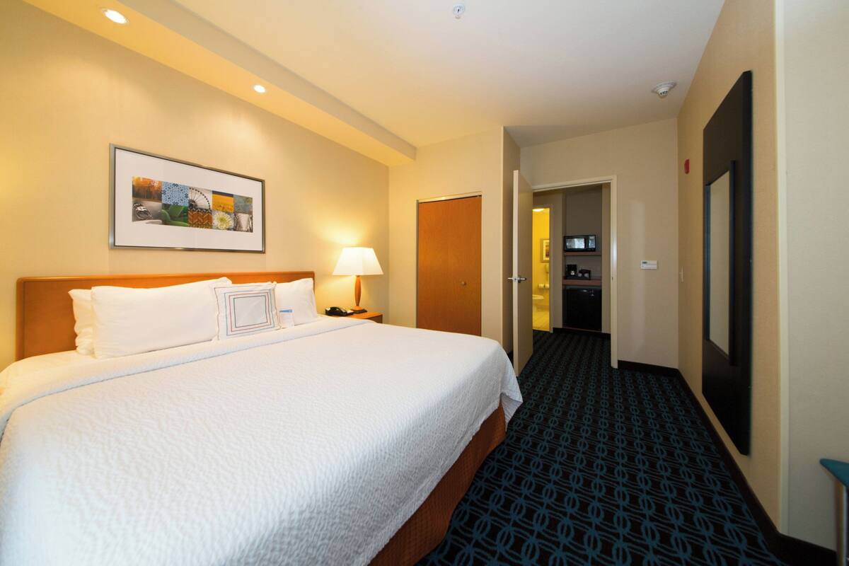 A hotel room with a large bed, a bedside lamp, a framed picture on the wall, and a carpeted hallway.