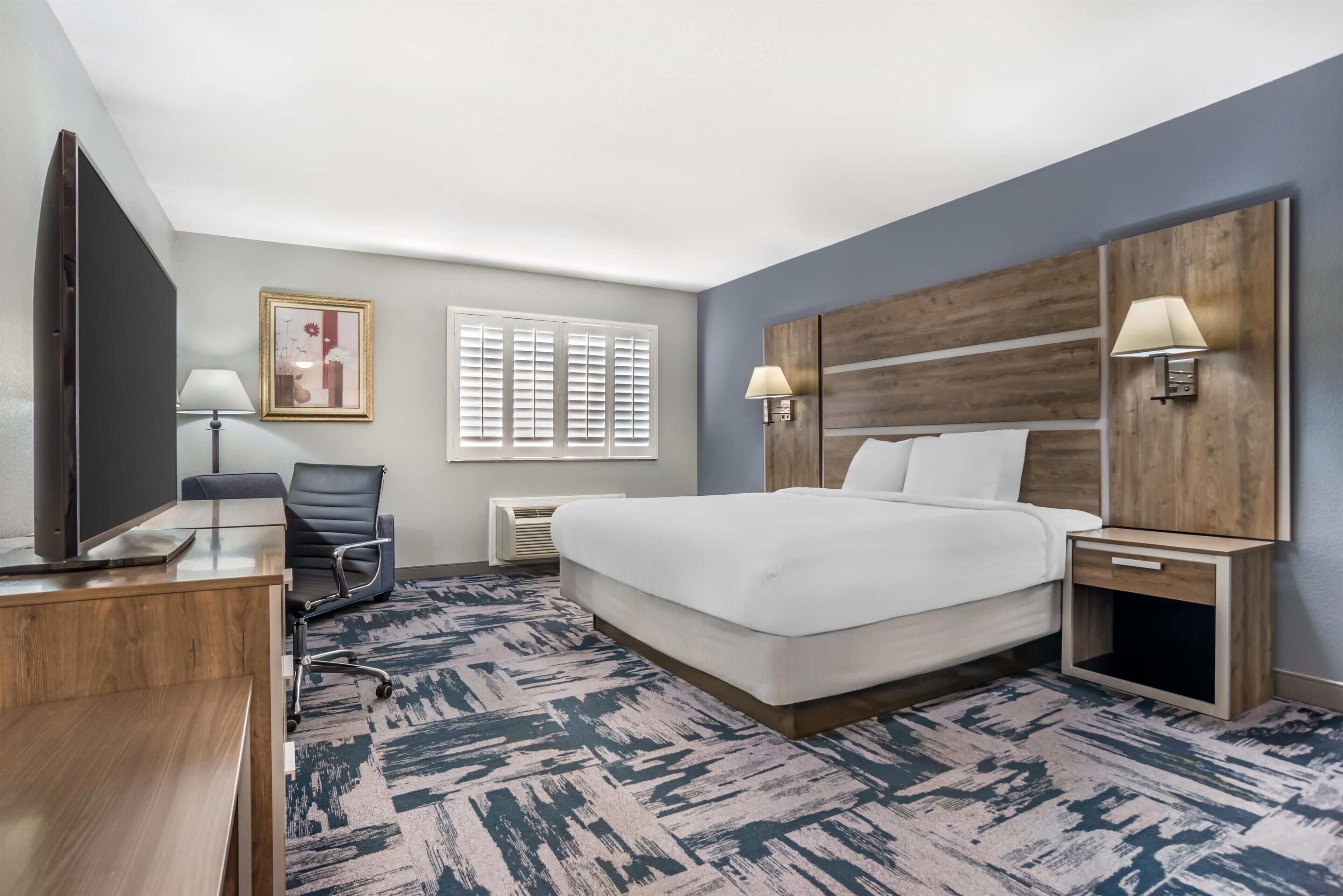 A hotel room with a bed, a desk, a chair, a TV, and a window with blinds.