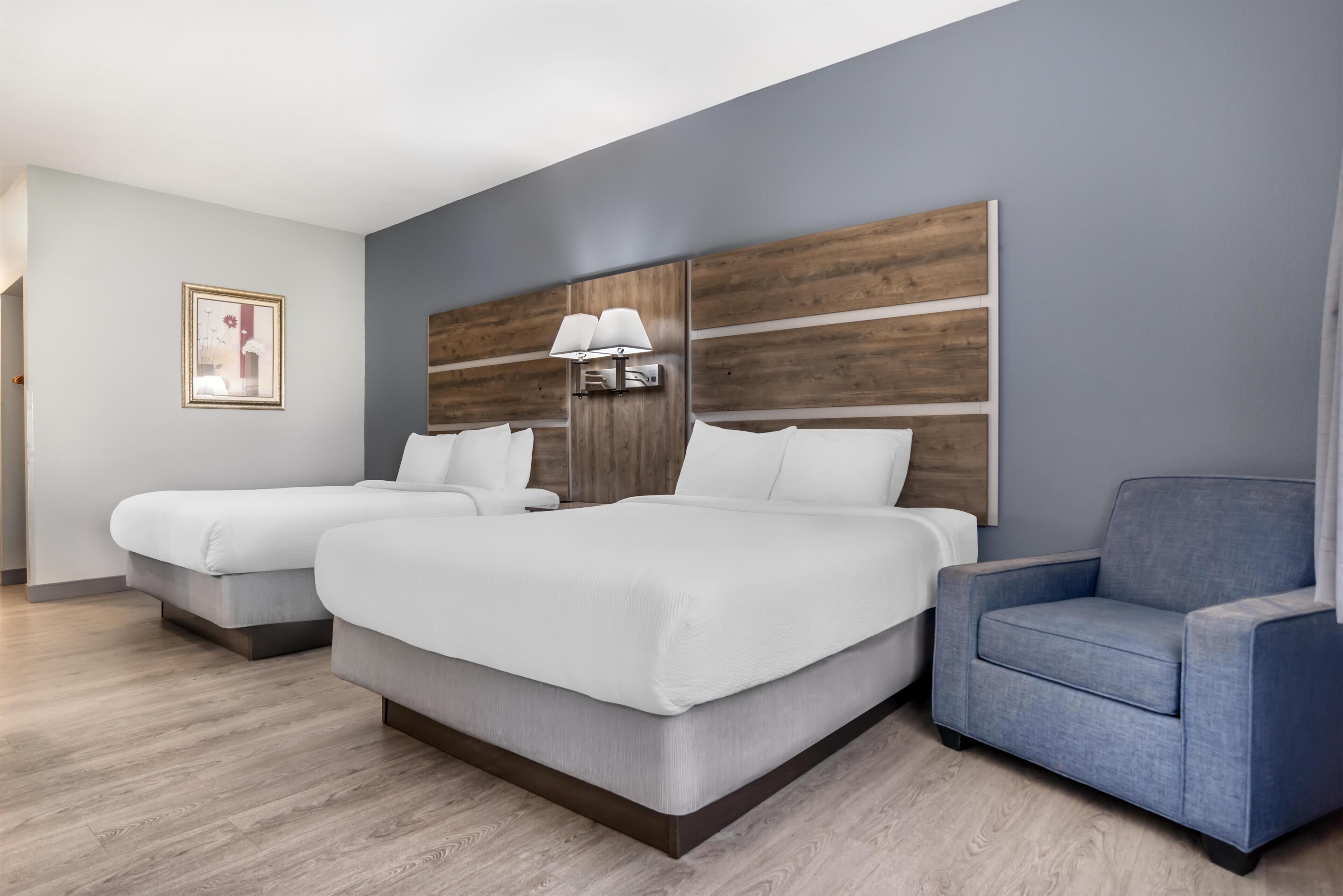 A hotel room with two beds, a blue armchair, a wooden headboard, and a framed picture on the wall.