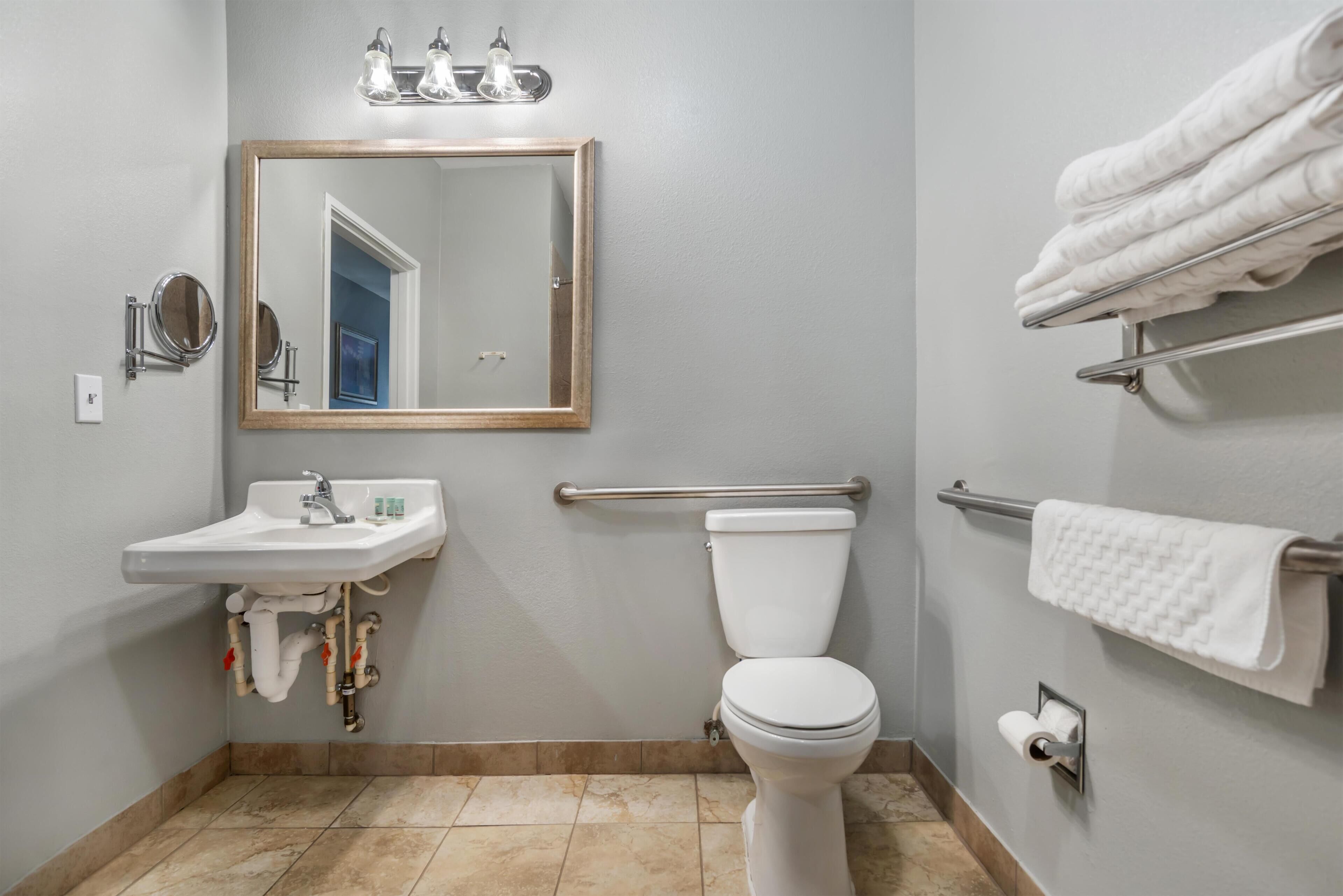 Un baño con lavamanos, espejo, inodoro, toallero y barras de apoyo.