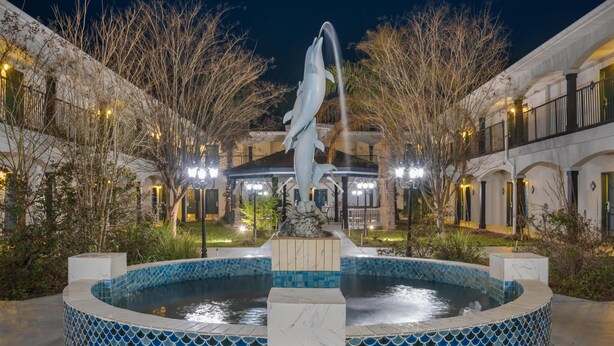 Ein Brunnen mit einer Delfinstatue in einem Innenhof, umgeben von Gebäuden mit Bogenfenstern und einem Pool mit blauen Mosaikfliesen.