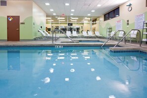 Uma piscina interior com superfície de água límpida, uma porta com um aviso e uma parede com relógio e cartazes informativos.