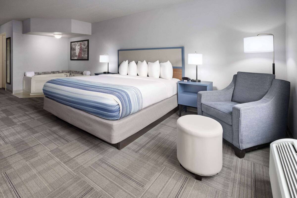 A hotel room with a large bed, a gray armchair, a white ottoman, a side table with a lamp, and a painting on the wall.