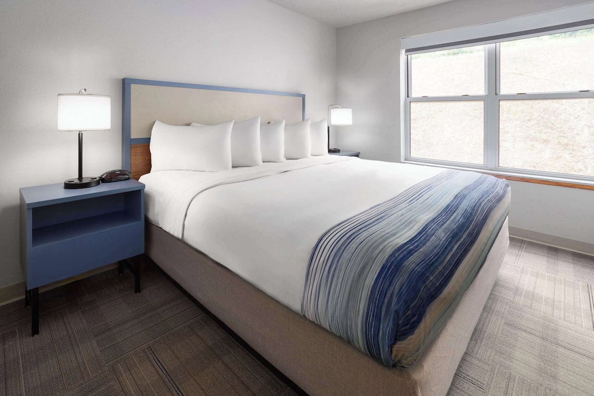 A hotel room with a large bed, two bedside tables with lamps, a window with a view, and a blue headboard.