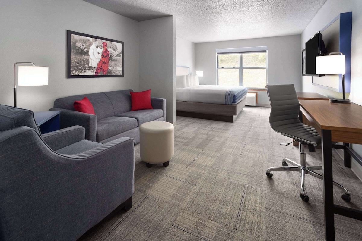 A hotel room with a grey sofa, a red pillow, a small ottoman, a bed, a desk with a chair, a TV, and a window.