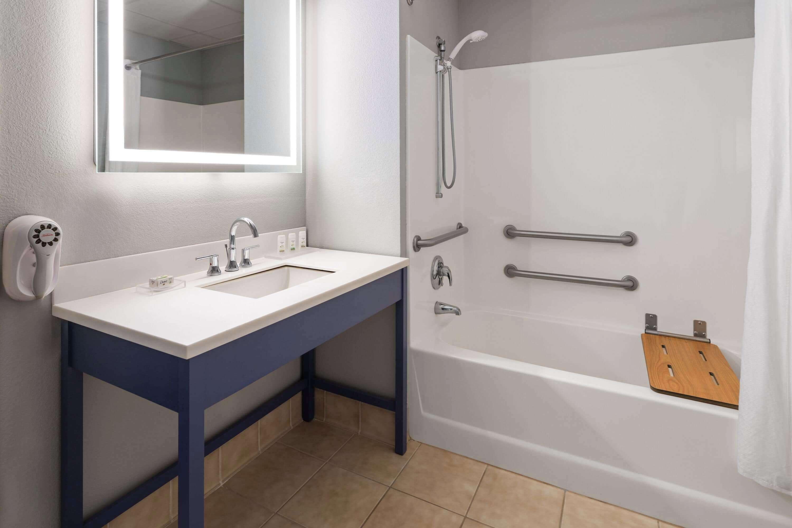 Un baño con lavamanos blanco, espejo, ducha con barras de apoyo y estante de madera.