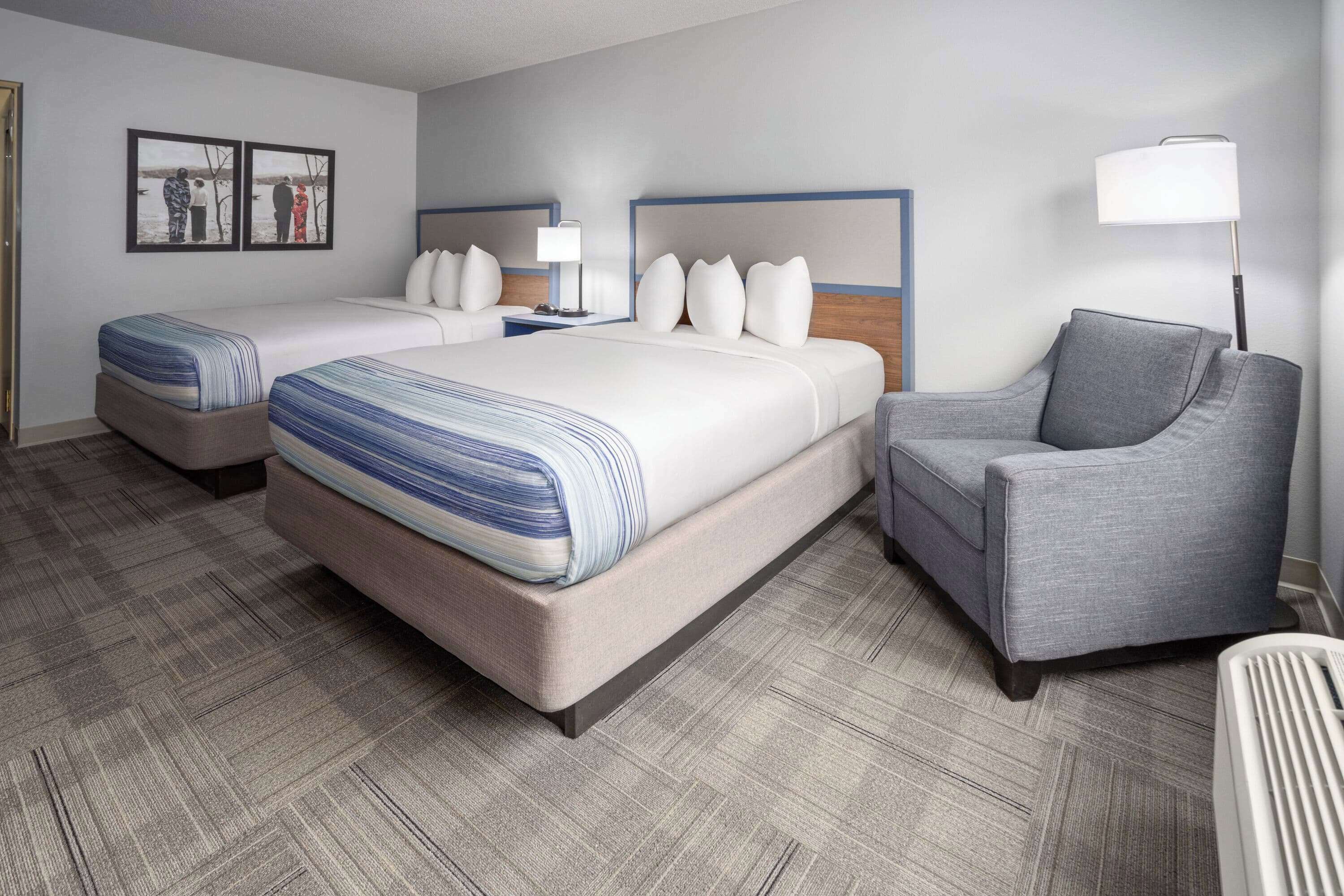 Habitación de hotel con dos camas, un sillón gris, una lámpara de pie y un televisor instalado en la pared.