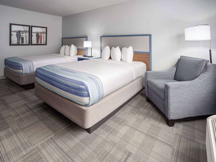 Habitación de hotel con dos camas, un sillón gris, una lámpara de pie y un televisor instalado en la pared.