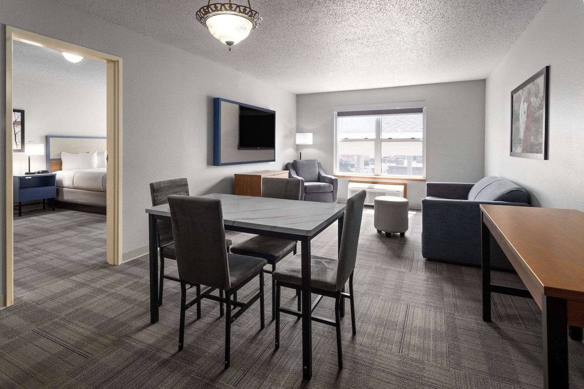 A modern hotel room with a dining area, a large window, and a flat-screen TV mounted on the wall.
