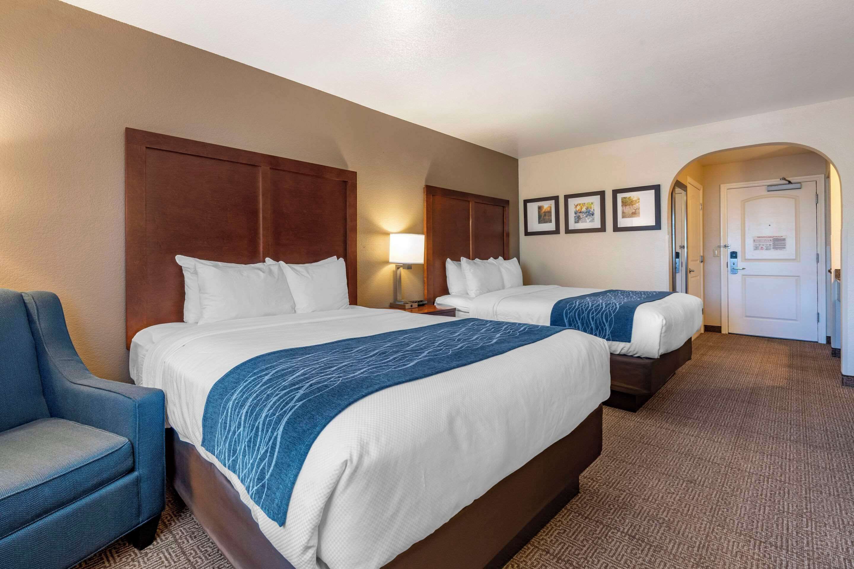 A hotel room with two beds, a blue armchair, a lamp, and framed pictures on the wall.
