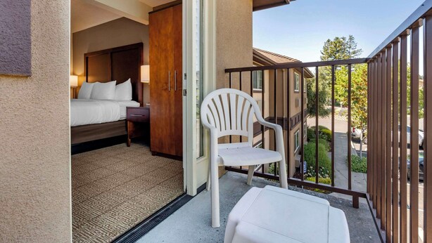 Un balcón con una silla blanca, una mesita y una cama con cabecera de madera.