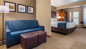 Habitación de hotel con un sofá azul, un reposapiés marrón, una cama con cabecera de madera y una ventana con persianas.