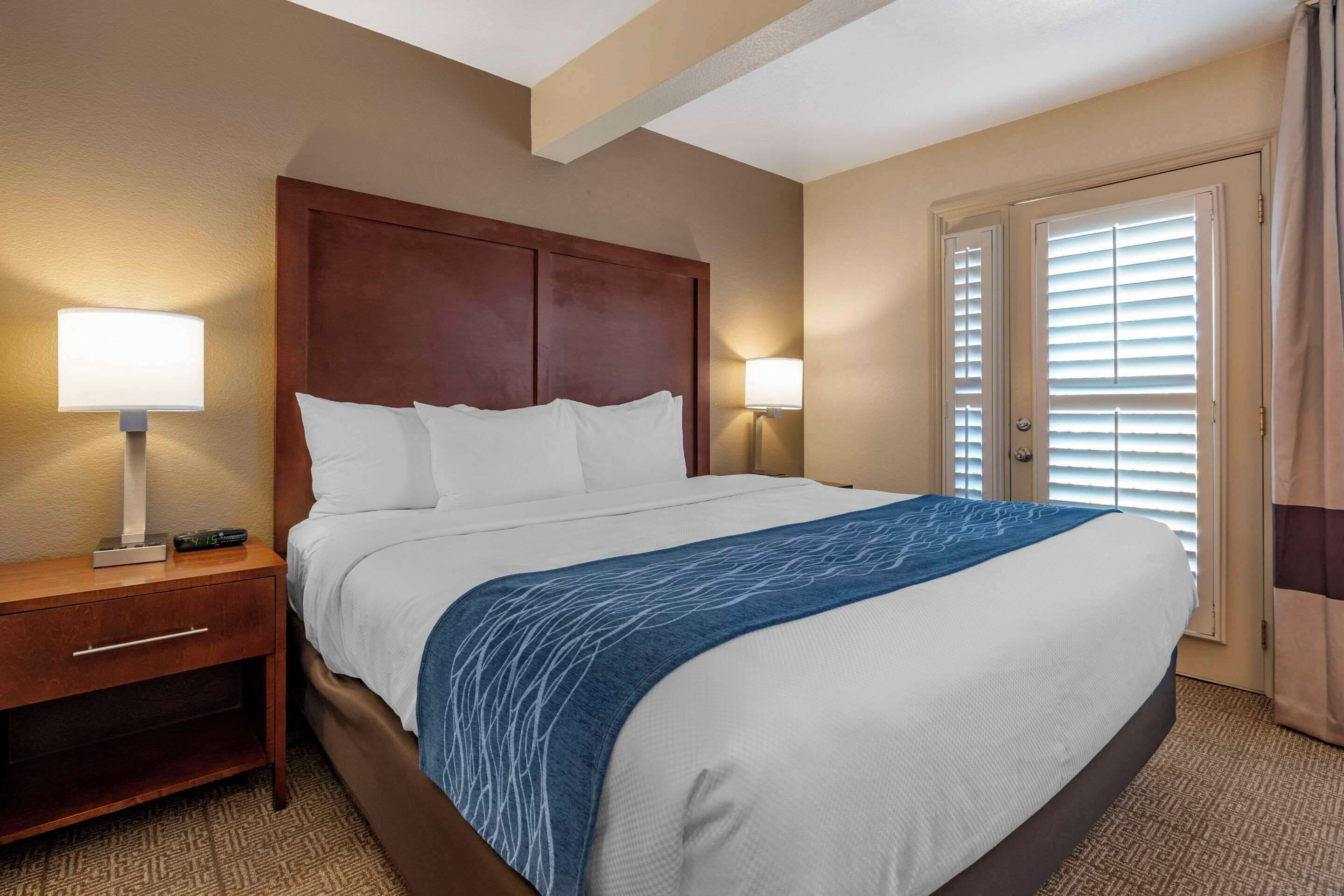 A hotel room with a large bed, wooden headboard, two bedside lamps, a nightstand, and a door with blinds.