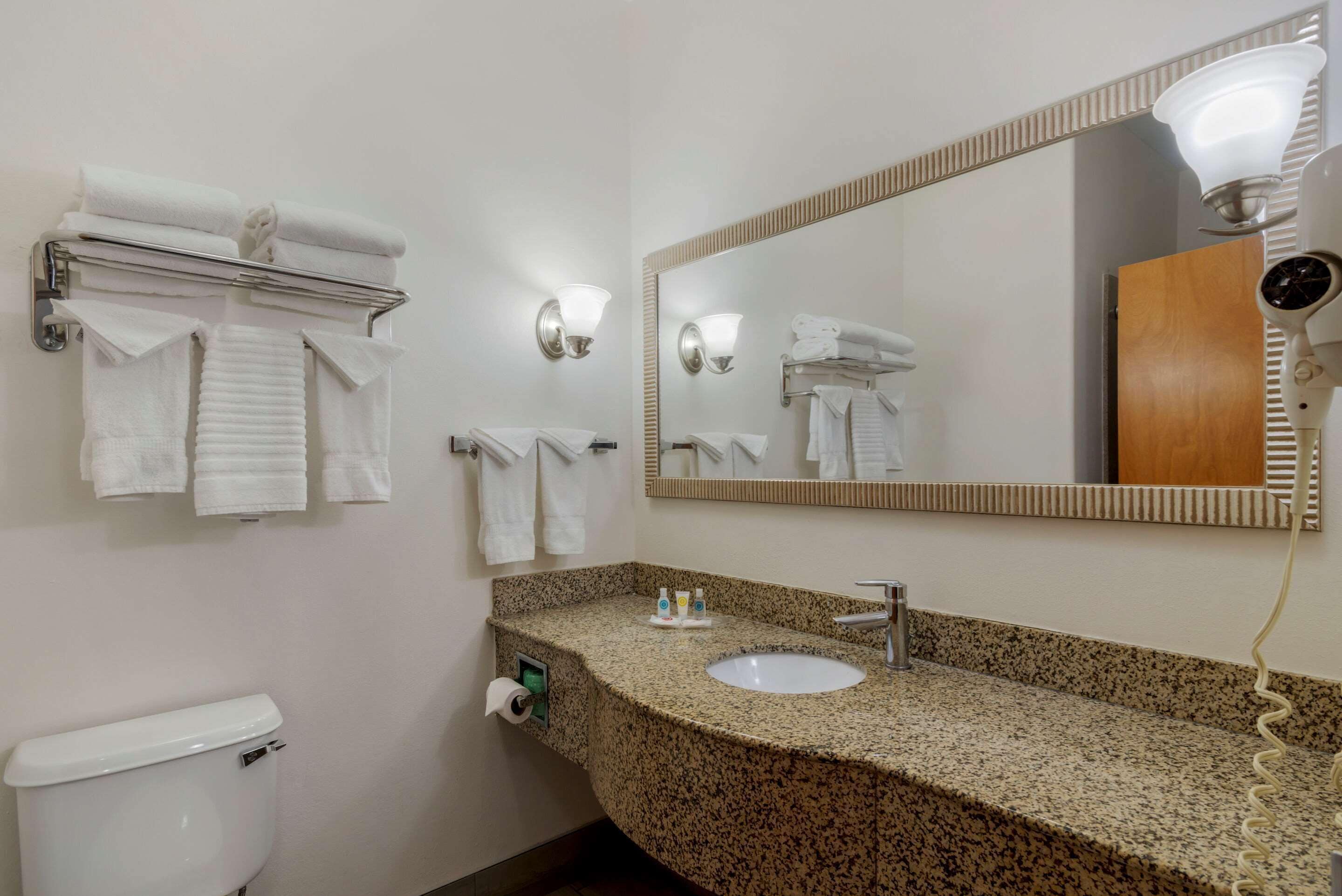 A hotel bathroom with a granite countertop sink, a mirror, a towel rack, and a hairdryer.