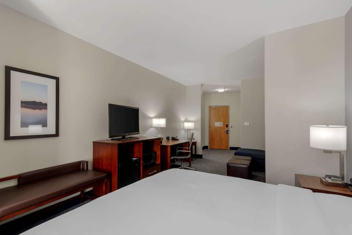 A hotel room with a large bed, a desk with a computer, a TV, a bench, and a door leading to another room.