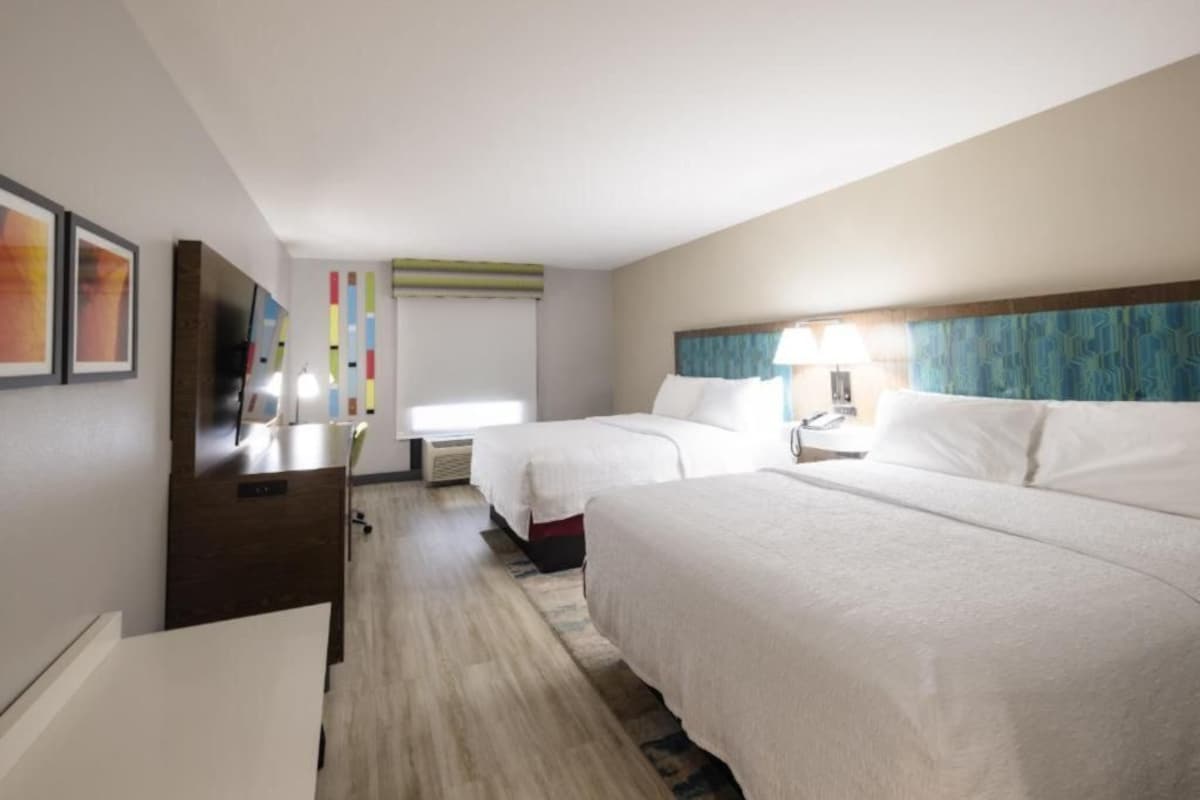 A hotel room with two beds, a desk, a TV, and a window with blinds.