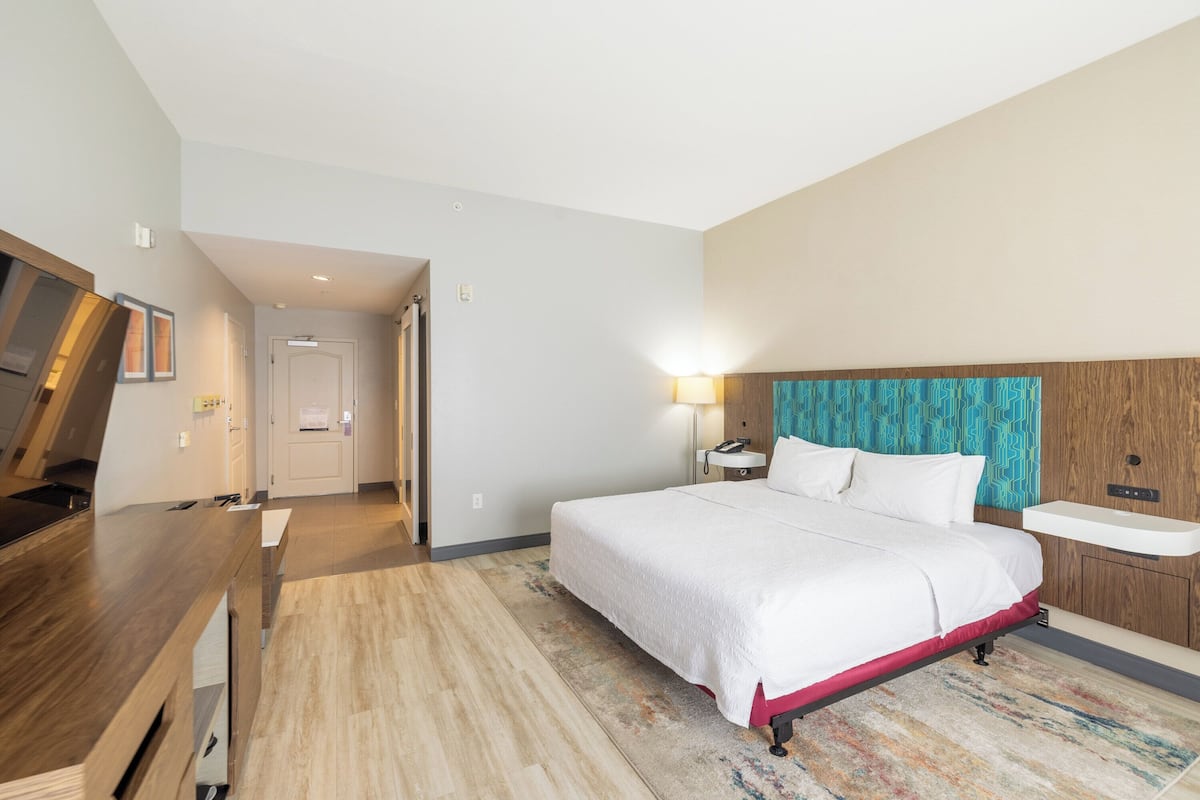A hotel room with a bed, bedside table, and a desk with a chair. There is a bathroom with a sink and mirror.