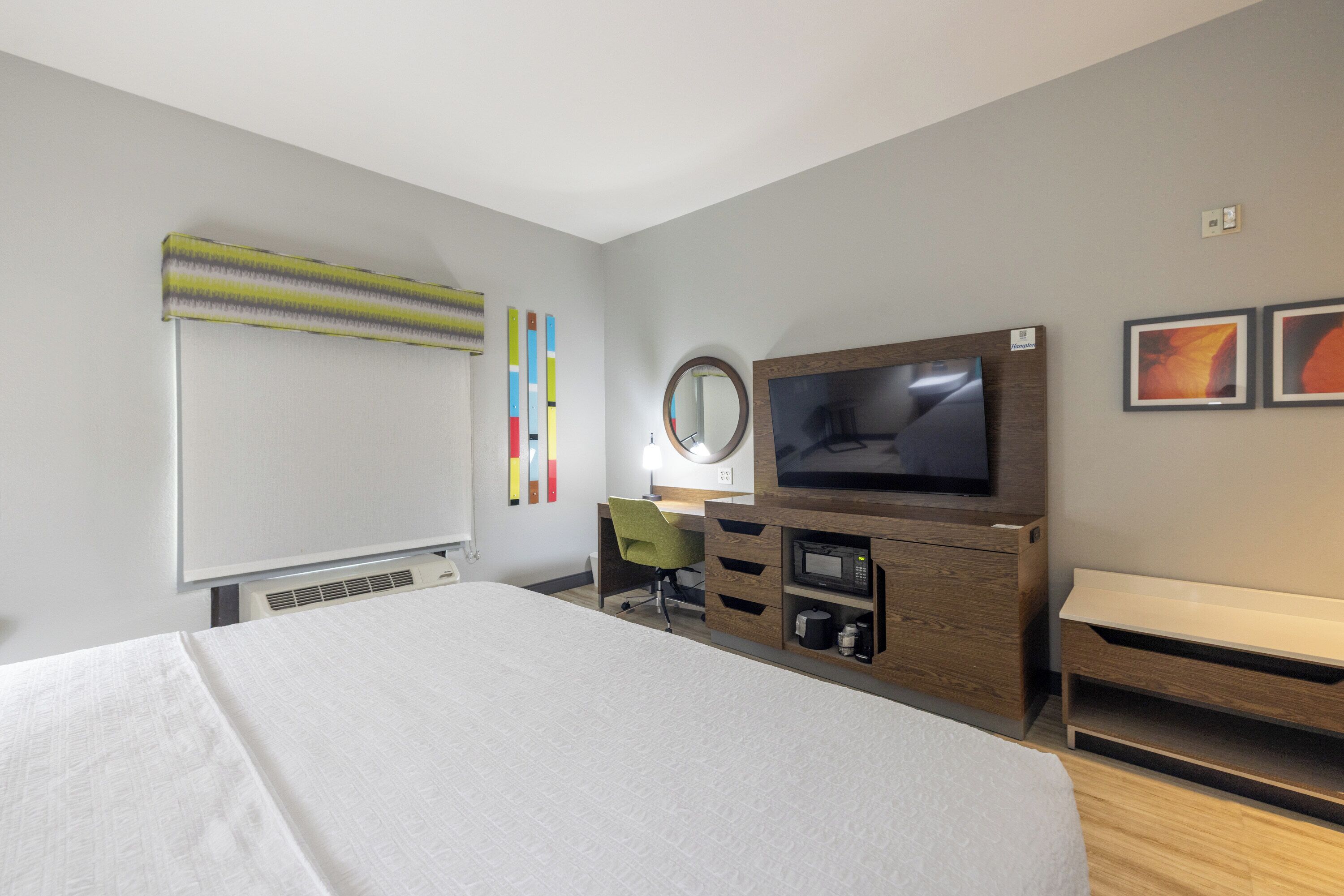 A hotel room with a large bed, a desk with a chair, a television, a microwave, and a small shelf.