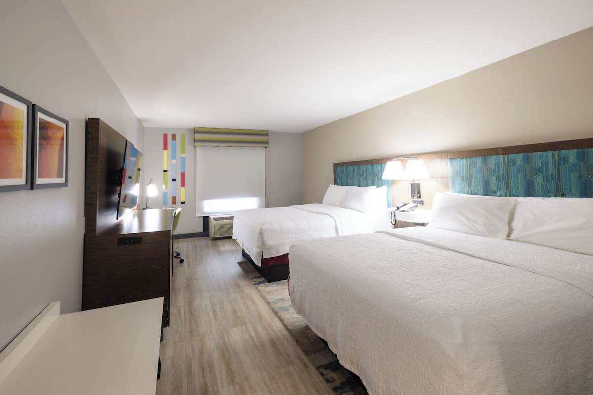 A hotel room with two beds, a desk, a TV, and a window with blinds.