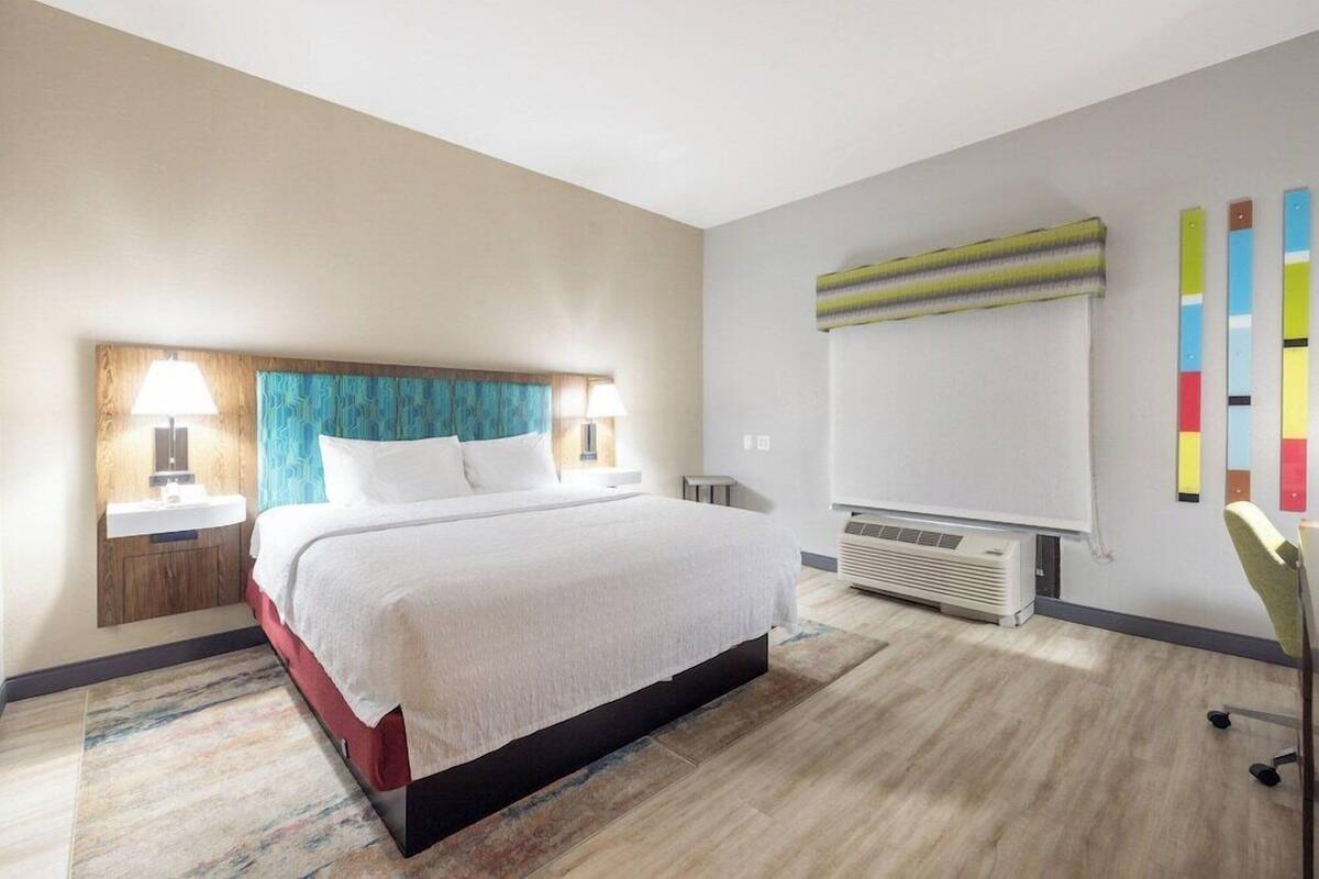 Une chambre d’hôtel avec un lit, des lampes de chevet, un climatiseur fixé au mur et une œuvre d’art abstraite aux couleurs vives.
