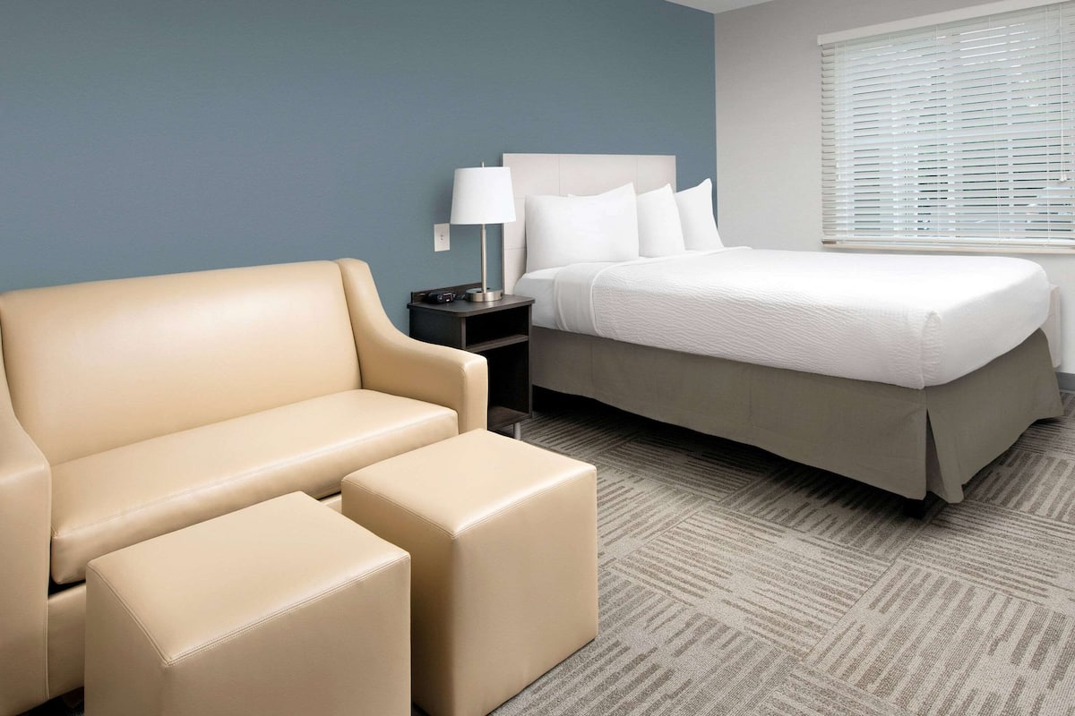 Una habitación de hotel con un sofá color beige, un reposapiés a juego, una cama con ropa blanca, una mesita de noche con lámpara y una ventana con persianas.