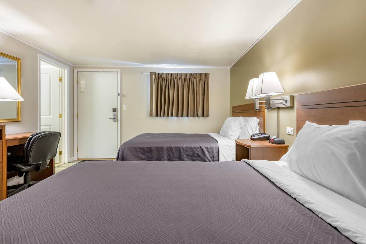A hotel room with two beds, a desk with a chair, a bedside table with a lamp, and a door.