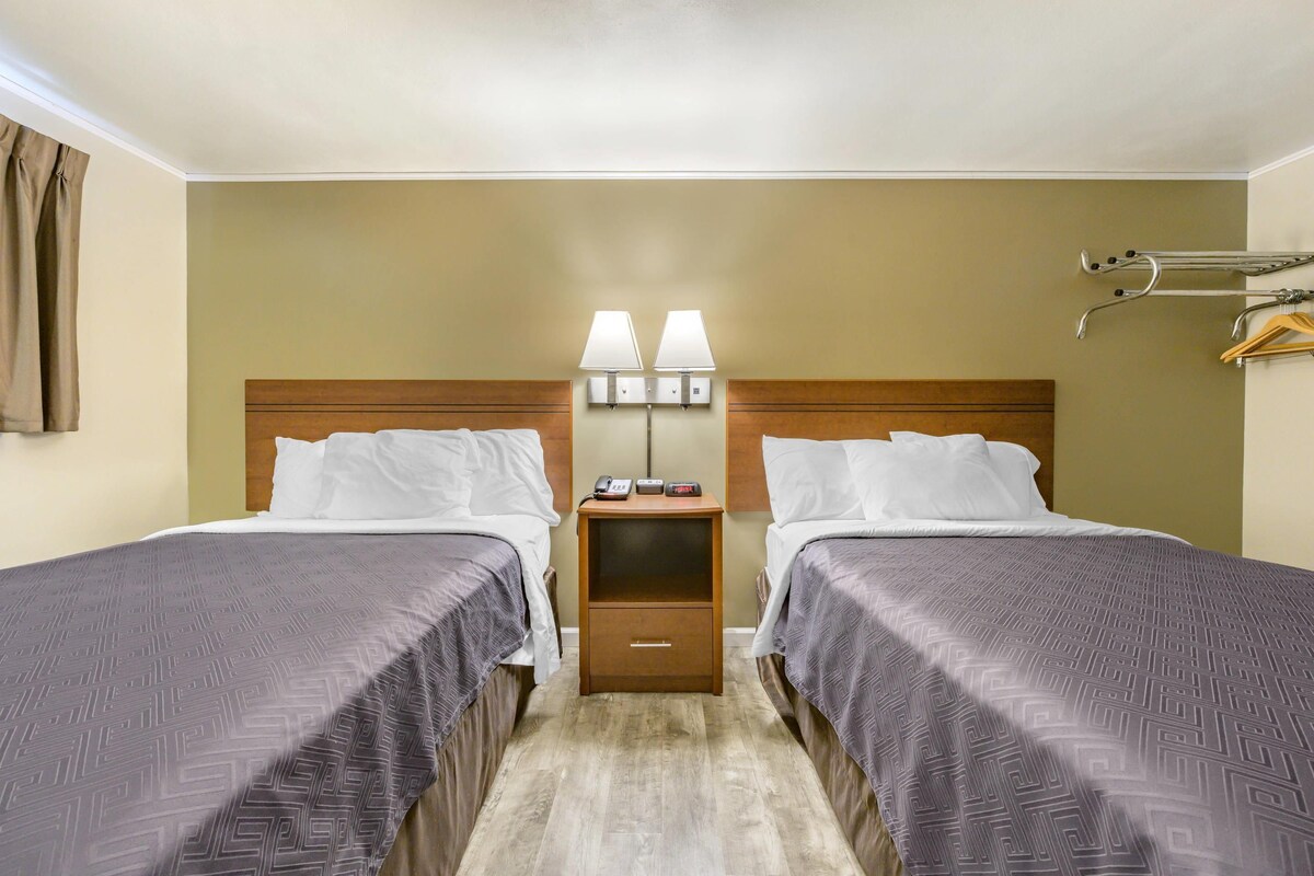 A hotel room with two beds, a nightstand with lamps, a small drawer, and a coat rack.