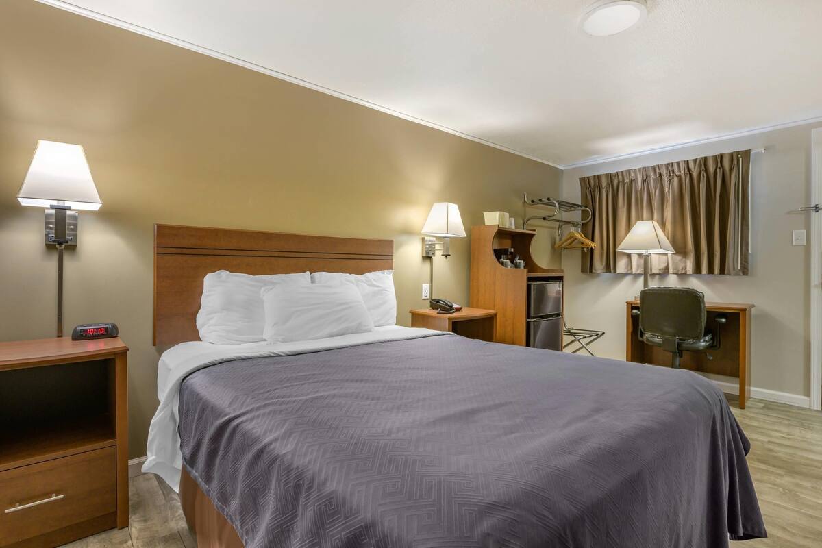 A hotel room with a bed, bedside lamps, a desk with a chair, a mirror, and a coat rack.