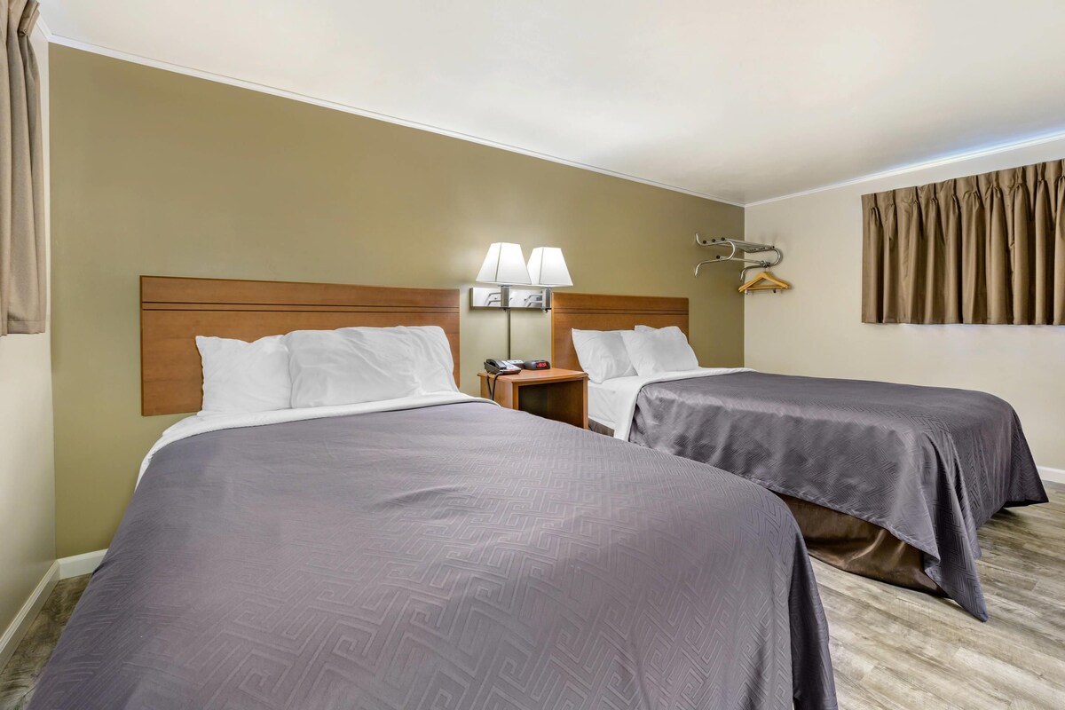 A hotel room with two beds, a nightstand with a lamp, a coat hanger, and a window with curtains.