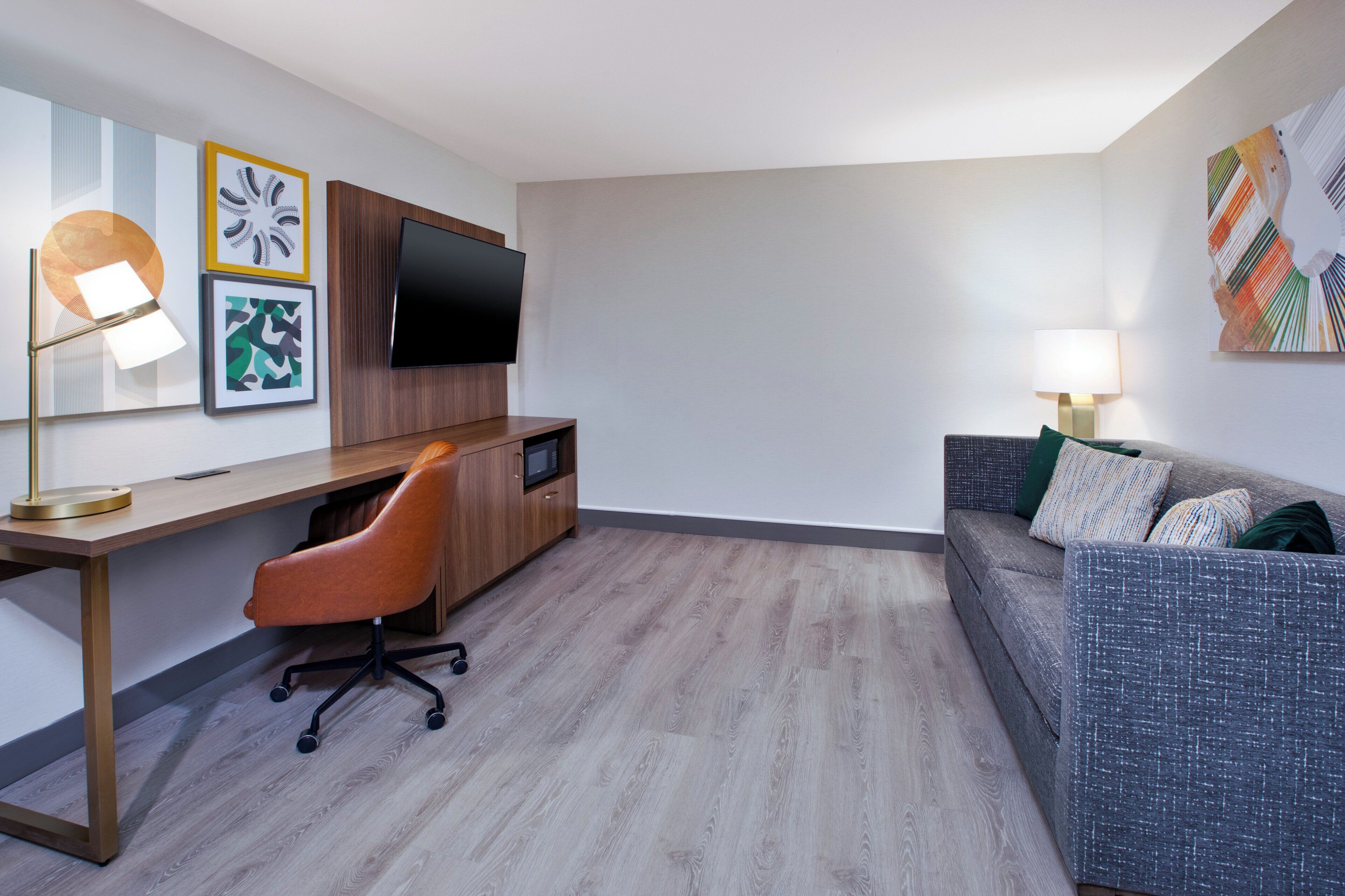 A modern hotel room with a wooden desk, a comfortable sofa, a flat-screen TV, and artwork on the walls.