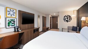 A hotel room with a large bed, a desk with a chair, a TV mounted on the wall, and a small table with a lamp.