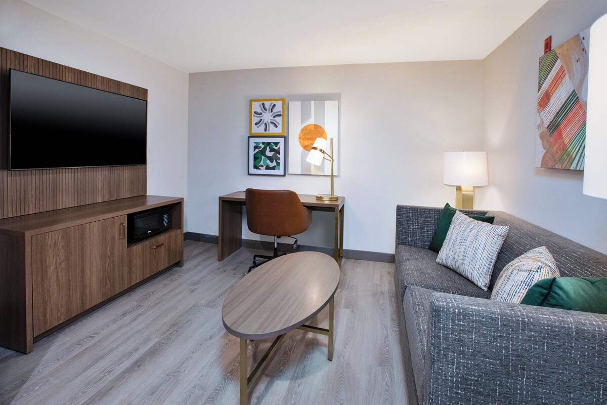A modern living room with a flat-screen TV, a wooden cabinet, a desk with a chair, a grey sofa with patterned cushions, and artwork on the walls.