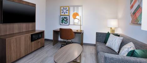 A modern living room with a flat-screen TV, a wooden cabinet, a desk with a chair, a grey sofa with patterned cushions, and artwork on the walls.
