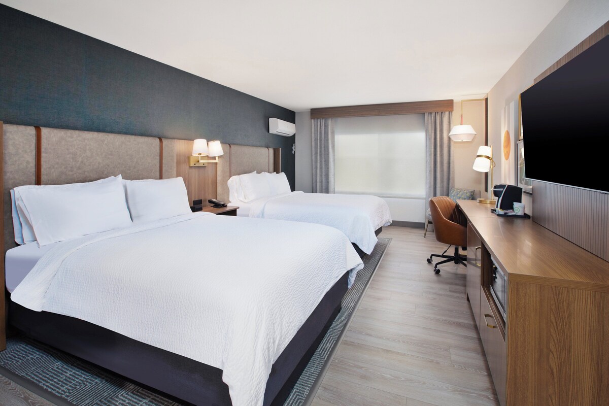A hotel room with two beds, a desk, a chair, a TV, and a large window.
