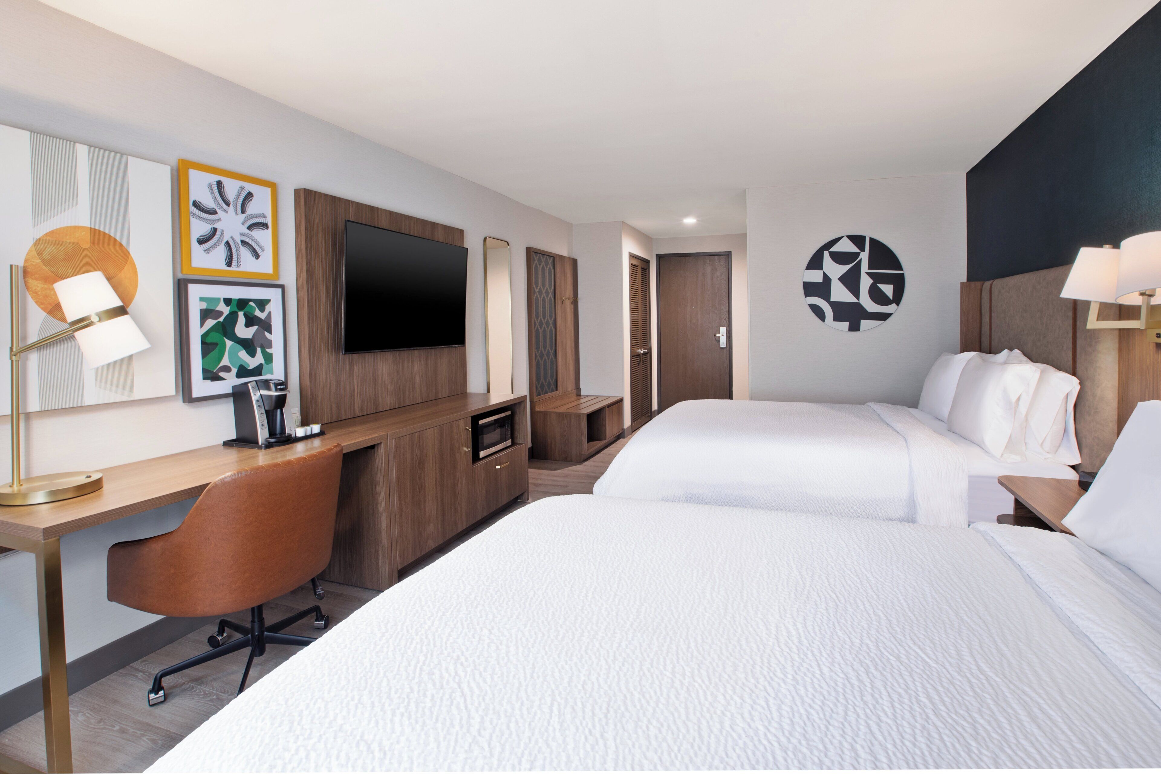 A hotel room with two beds, a desk, a chair, a TV, and artwork on the walls.
