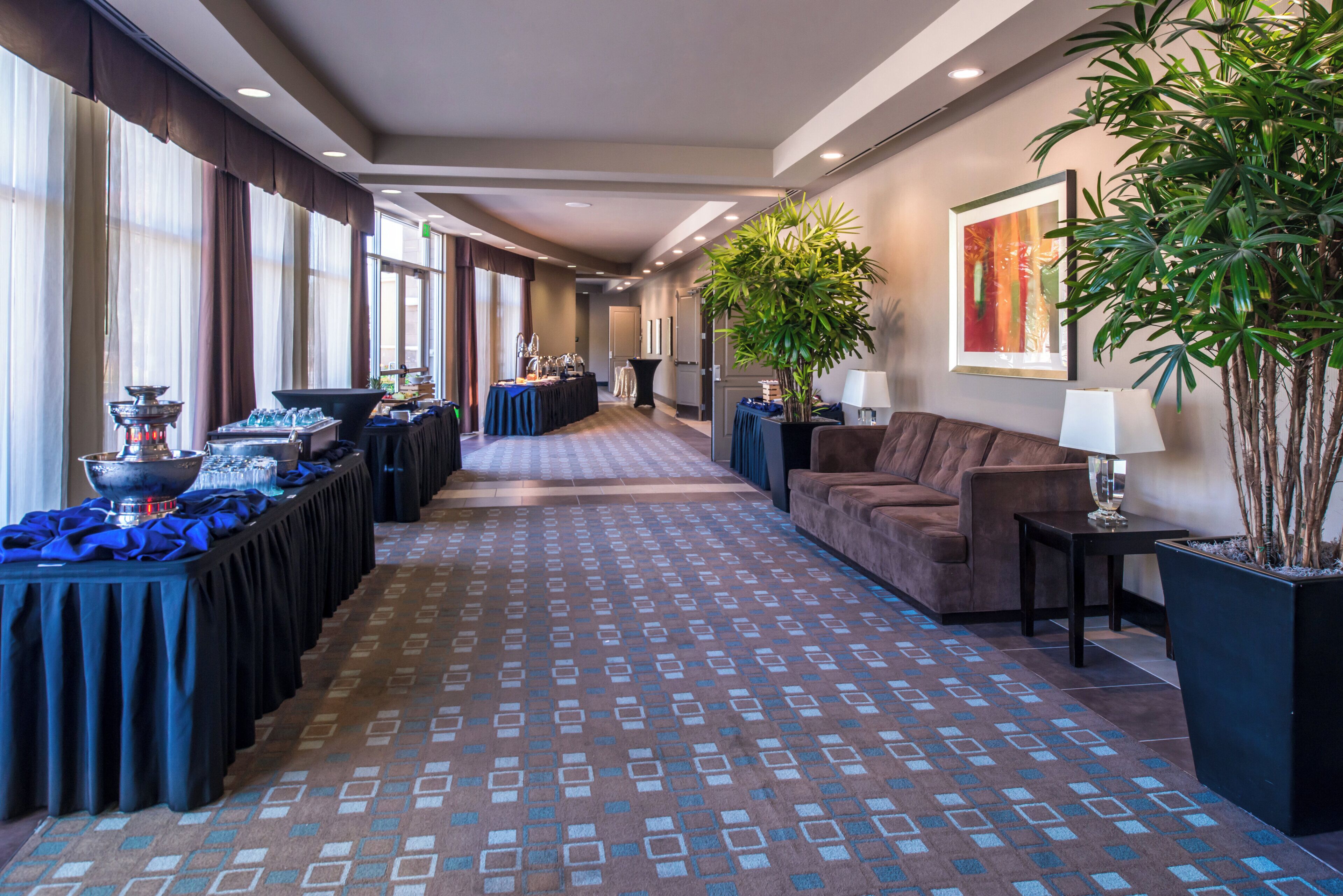 A hotel corridor with a patterned carpet, a reception desk, a sofa, and a large potted plant.