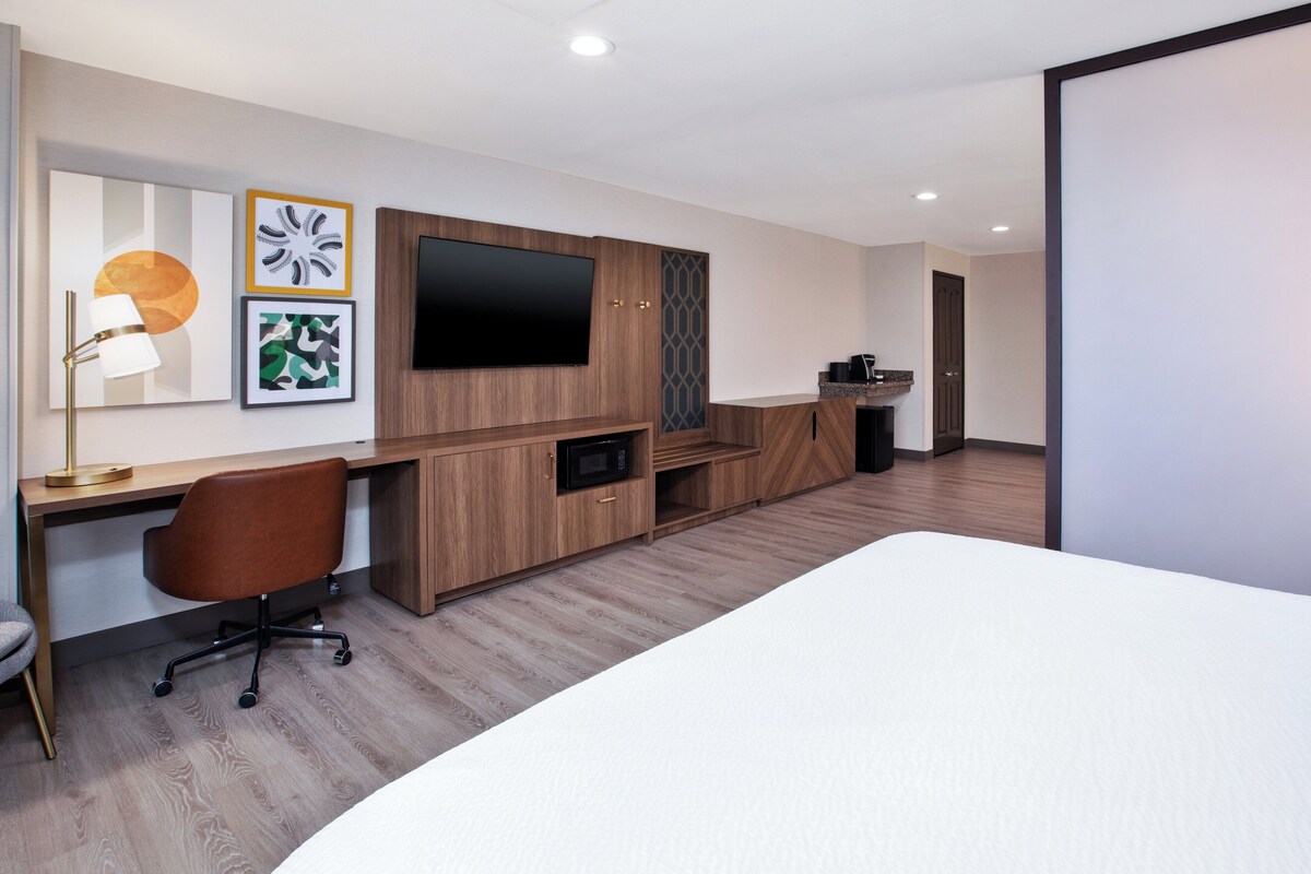 A modern hotel room with a wooden desk, a flat-screen TV, a bed, and artwork on the wall.