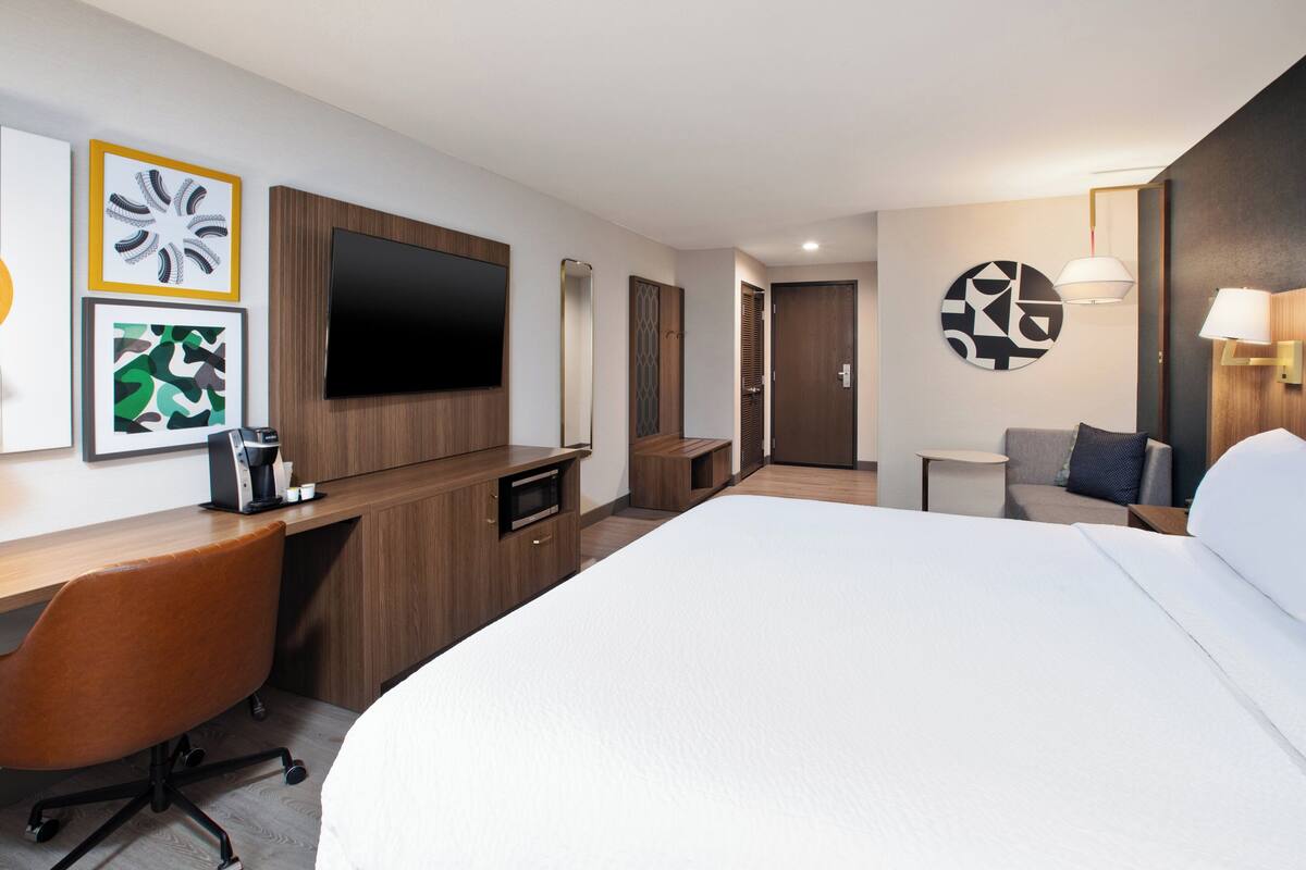 A hotel room with a large bed, a desk with a chair, a TV mounted on the wall, and a small table with a lamp.