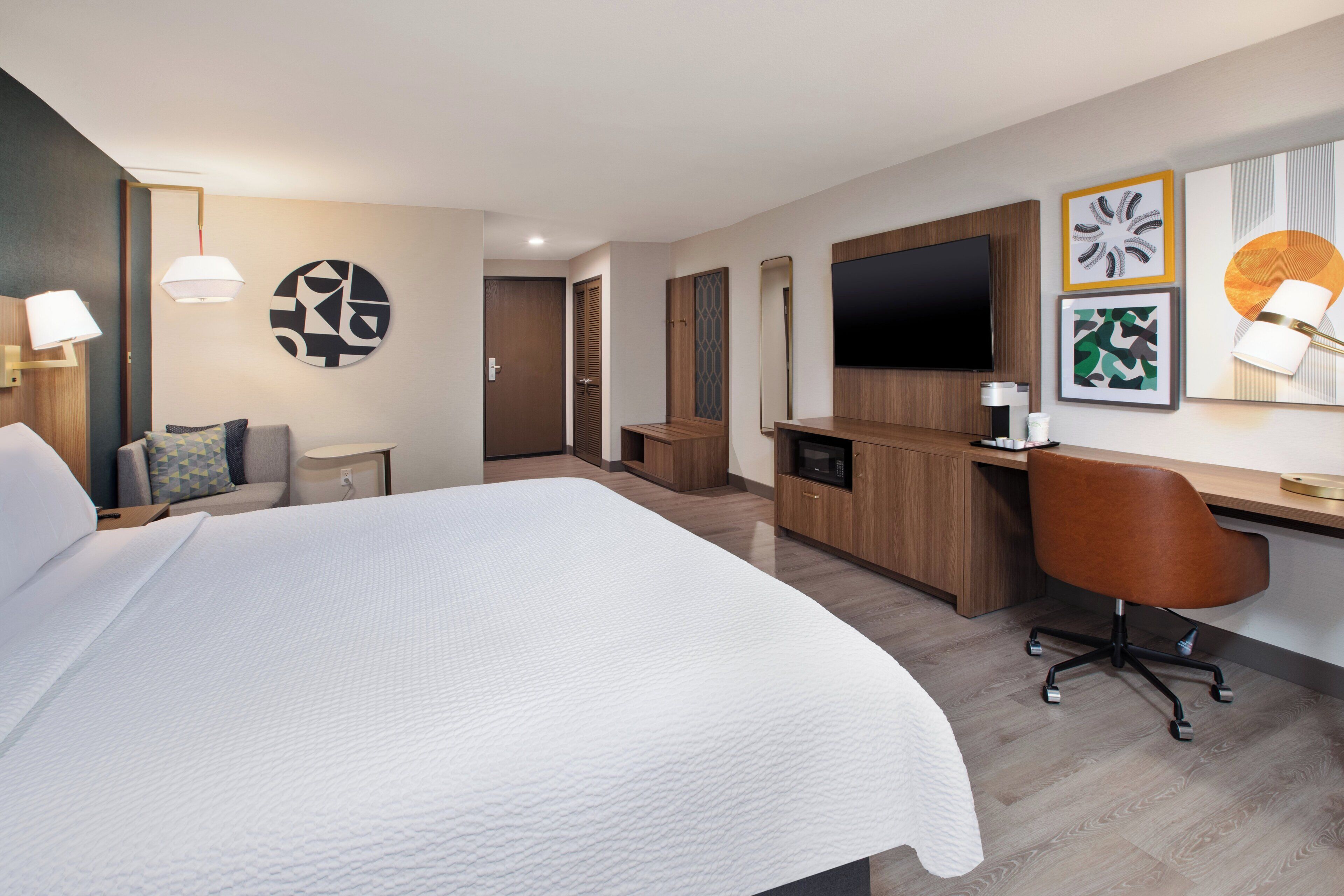 A hotel room with a large bed, a desk with a chair, a TV, and artwork on the walls.