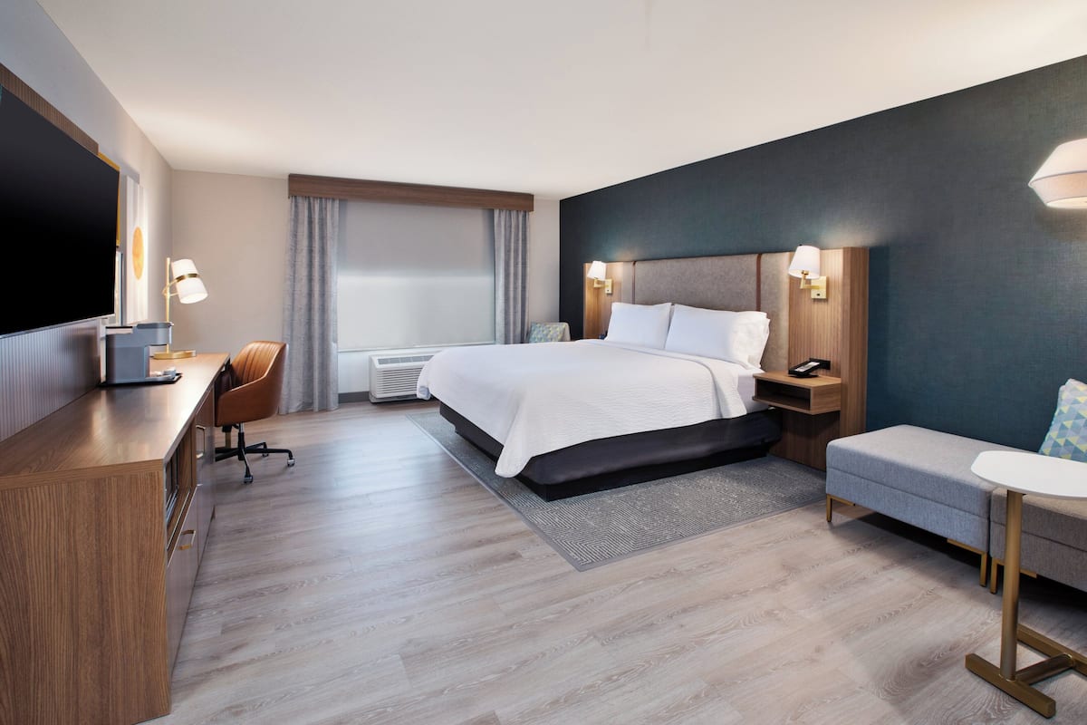 A modern hotel room with a large bed, a desk, a chair, a small table, and a TV.