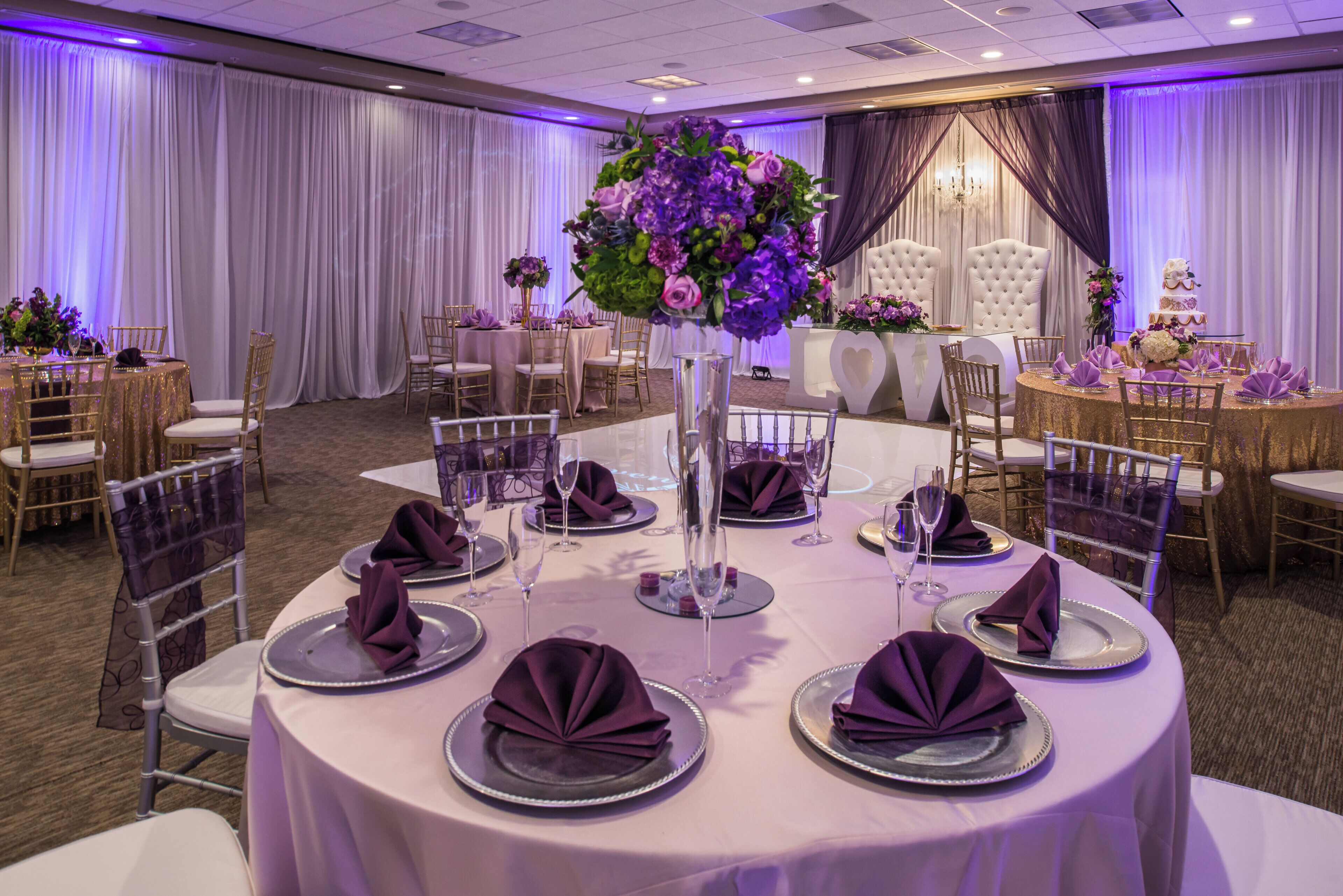 A banquet hall with round tables set for a formal event, featuring white tablecloths, purple napkins, and floral centerpieces.
